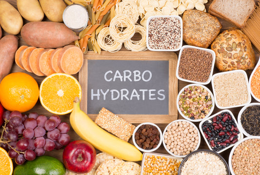 Various form of carbohydrates such as fruits, bread, pasta, and vegetables all around a sign that reads "Carbohydrates"